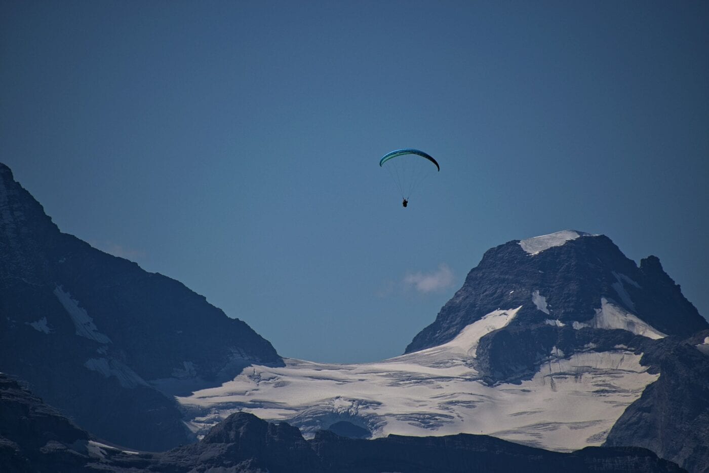 Paraglider flying above snow-capped mountain peaks under a clear blue sky